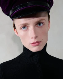 Close-up portrait of a model wearing a deep purple velvet cap and black turtleneck sweater against a plain neutral wall.