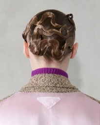 Rear view of a model with elaborate sculpted finger waves in brown hair wearing a tweed jacket over a purple collar for Prada F/W 24