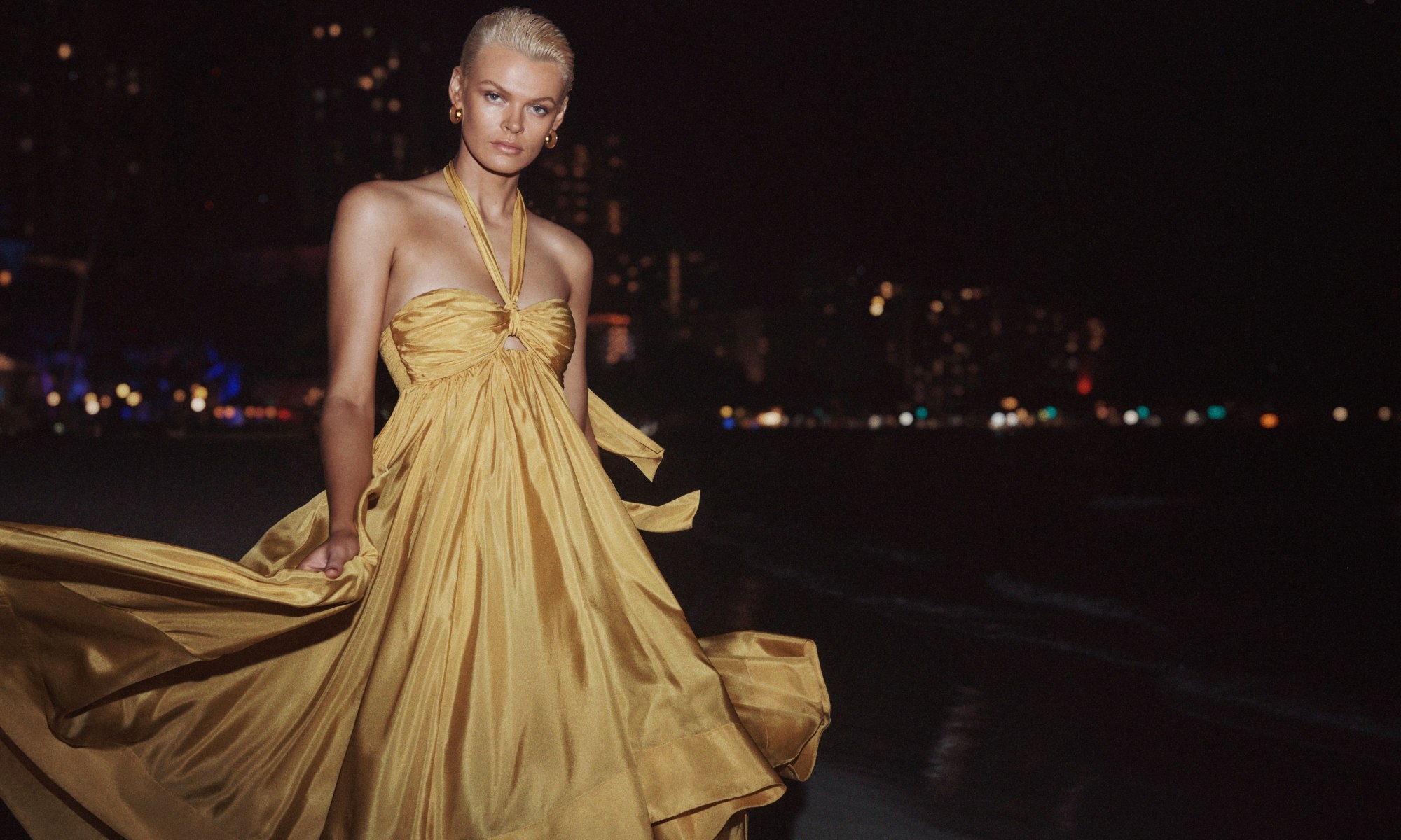Blonde woman in gold halter gown poses at the beach at night, city lights blurred in distance, windblown gown, fashion photography.