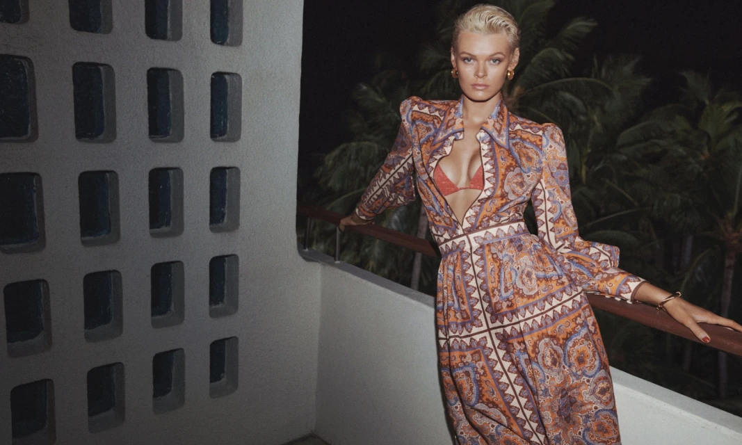 Attractive woman in a patterned dress stands on a balcony at night with palm trees in the background and architectural wall feature.