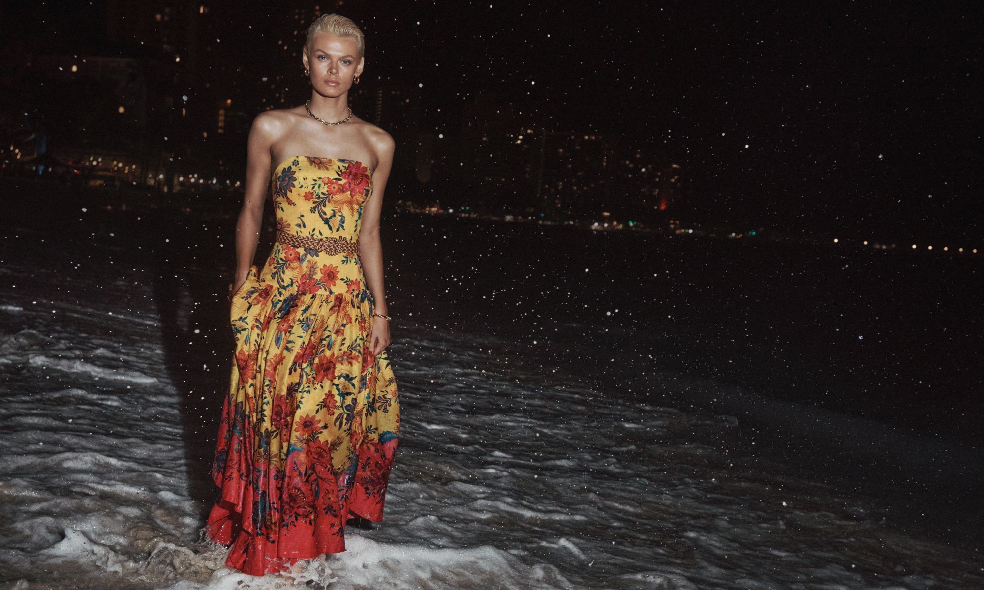 Woman in strapless floral dress stands on a dark, wet beach at night, with city lights visible across the water.