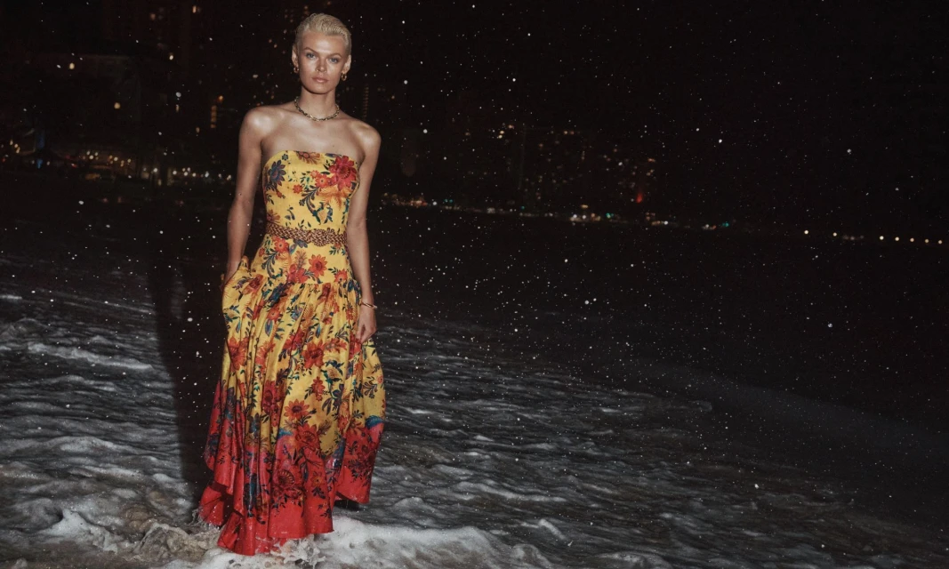 Woman in strapless floral dress stands on a dark, wet beach at night, with city lights visible across the water.