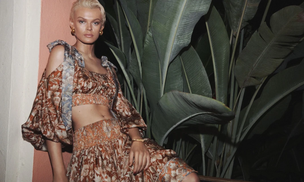 A blonde woman models a two-piece floral print outfit with shoulder cutouts in a tropical outdoor setting with large green leaves.