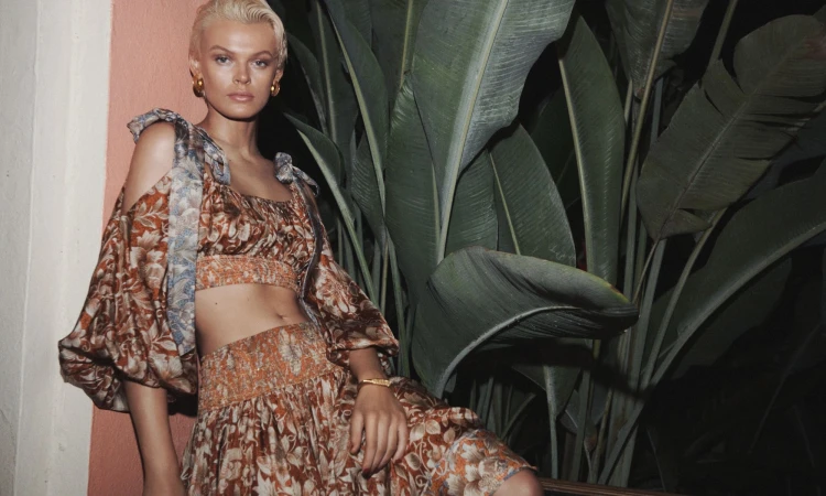 A blonde woman models a two-piece floral print outfit with shoulder cutouts in a tropical outdoor setting with large green leaves.