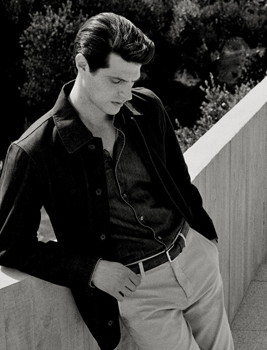 Black and white portrait of a man in a dark button-down shirt and jacket leaning against a concrete wall outdoors.
