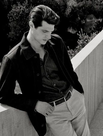 Black and white portrait of a man in a dark button-down shirt and jacket leaning against a concrete wall outdoors.