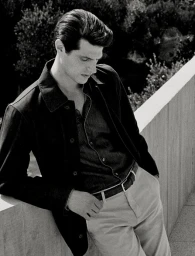 Black and white portrait of a man in a dark button-down shirt and jacket leaning against a concrete wall outdoors.