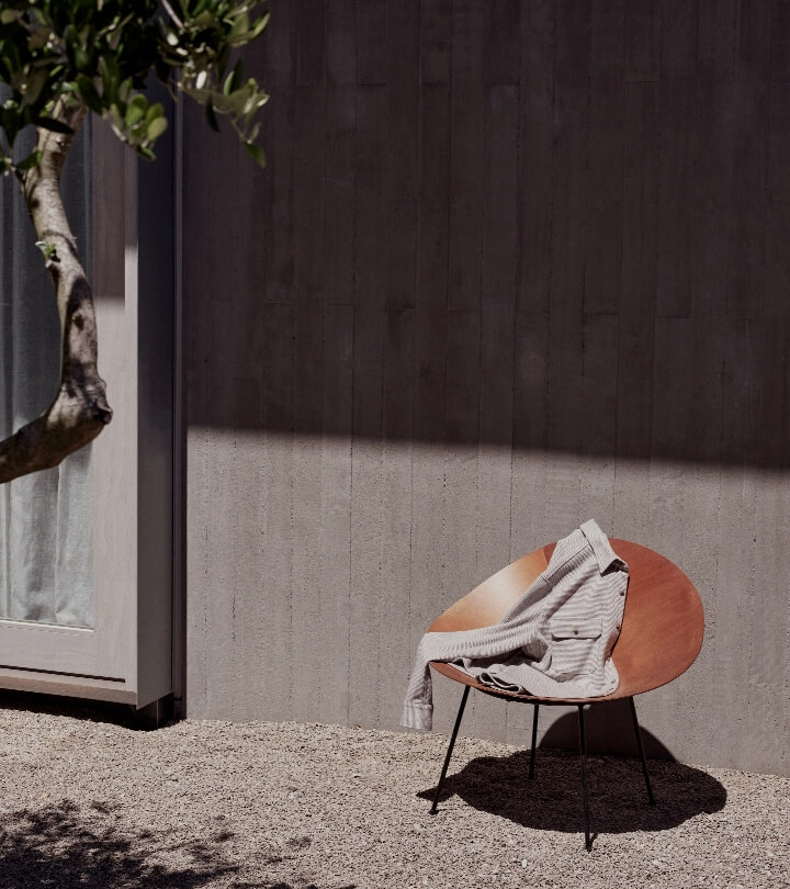 Minimalist shot of a wooden chair with a sweater on it, placed outdoors on a gravel surface with a grey wall and greenery in the background.