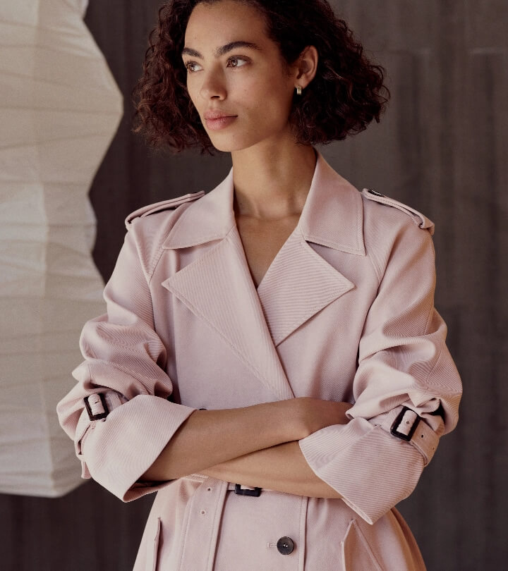Portrait of a woman wearing a pink trench coat with her arms crossed, standing indoors with a neutral expression, looking away from the camera.