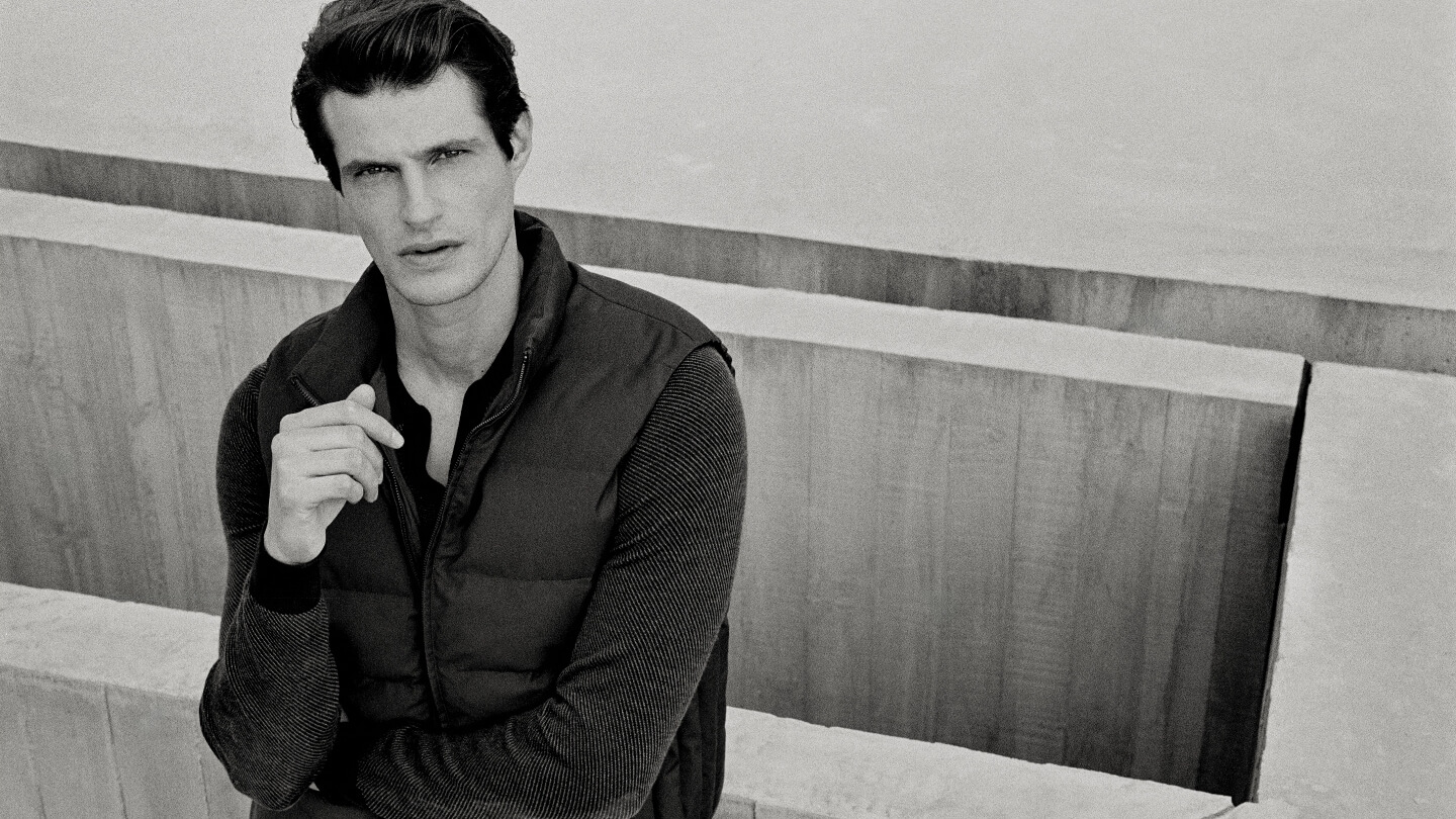Man wearing a puffer vest over a ribbed sweater against concrete architecture in a black and white portrait.