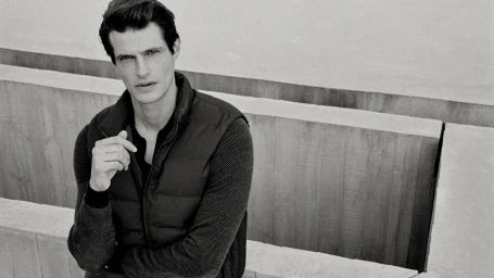 Man wearing a puffer vest over a ribbed sweater against concrete architecture in a black and white portrait.