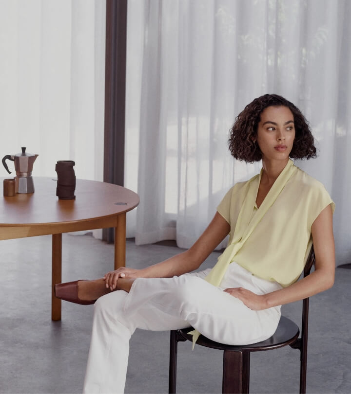 Adult female model poses indoors in a yellow blouse and white pants, seated near a wooden table with a coffee maker and cup.