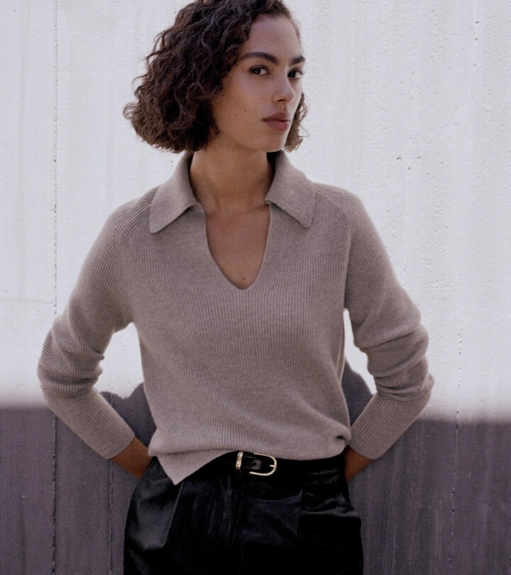A woman poses in a studio wearing a beige, collared, long-sleeved sweater and black leather pants with a black belt and silver buckle.
