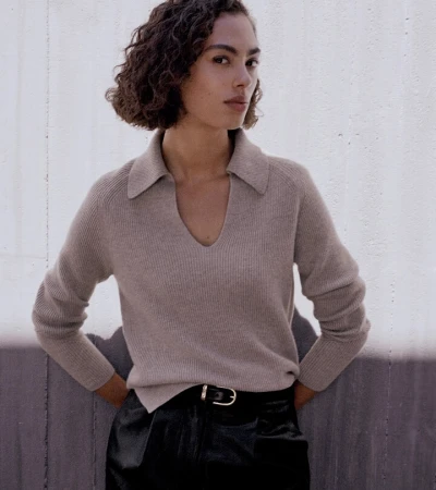 A woman poses in a studio wearing a beige, collared, long-sleeved sweater and black leather pants with a black belt and silver buckle.