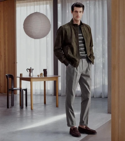 Man standing indoors wearing an olive suede bomber jacket, striped sweater, grey trousers, and brown loafers