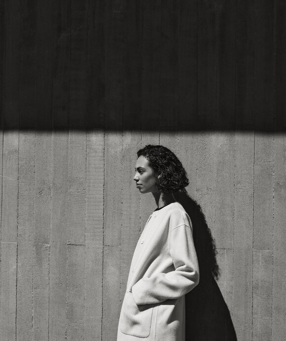 Model in profile wearing a light-coloured coat against a concrete wall with strong directional lighting and shadows.