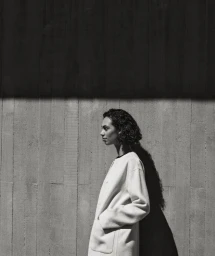Model in profile wearing a light-coloured coat against a concrete wall with strong directional lighting and shadows.
