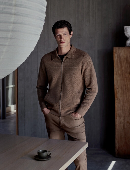 Stylish adult male in brown jacket and pants stands indoors with hands in pockets near a modern table and cup, posing in soft lighting.