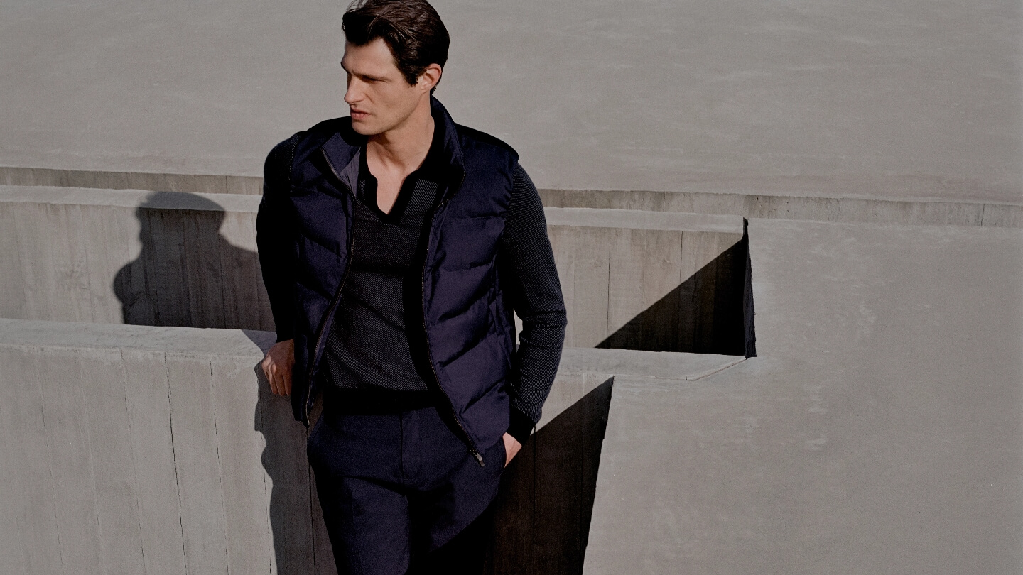 Adult male leaning against concrete structure wearing a dark puffer vest over a grey sweater, with concrete steps in background.