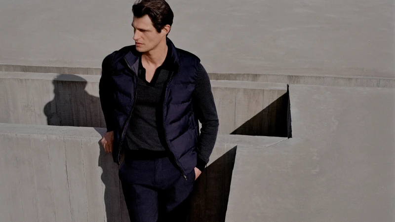 Adult male leaning against concrete structure wearing a dark puffer vest over a grey sweater, with concrete steps in background.