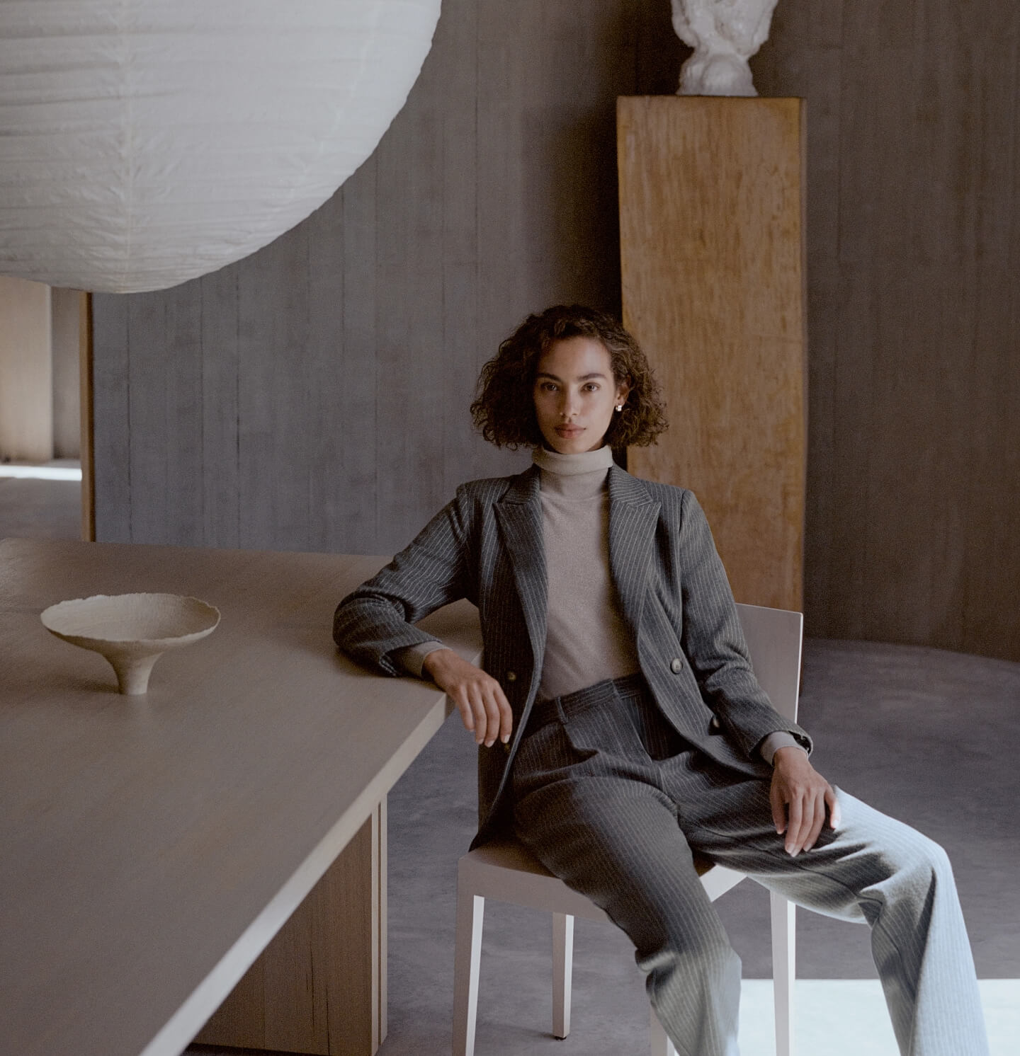 A stylish woman poses in a suit indoors, seated at a modern table with a sculpture and lighting fixture enhancing the minimalist scene.