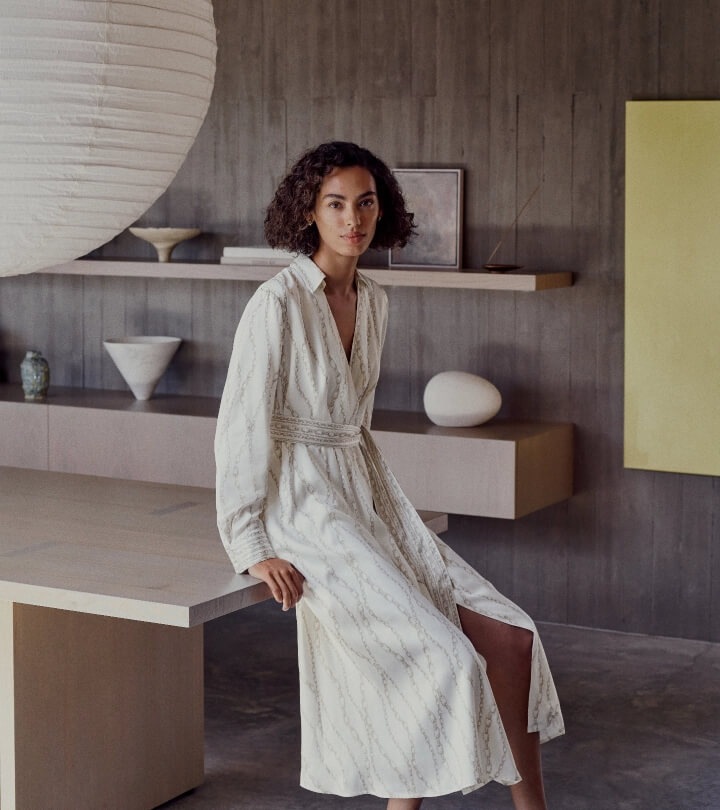 A brunette woman poses indoors, sitting on a table and wearing a white patterned dress. The backdrop includes shelves and a light fixture.