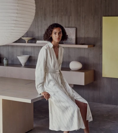 Model seated on a large wooden table in an elegant interior wearing a patterned cream midi dress
