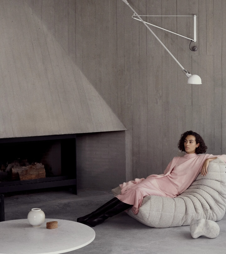 Woman in pink dress and black boots sits in an interior space with a fireplace and marble table, illuminated by a sleek white lamp.