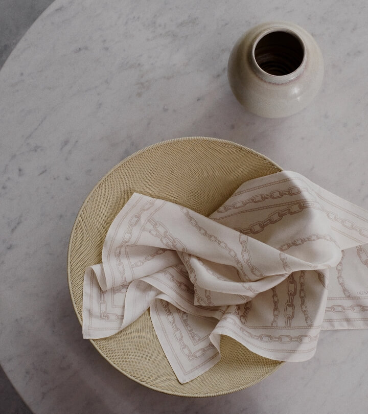 Still life featuring a beige woven plate with a patterned white cloth, a ceramic vase, and a marble surface, showcasing soft, natural lighting.