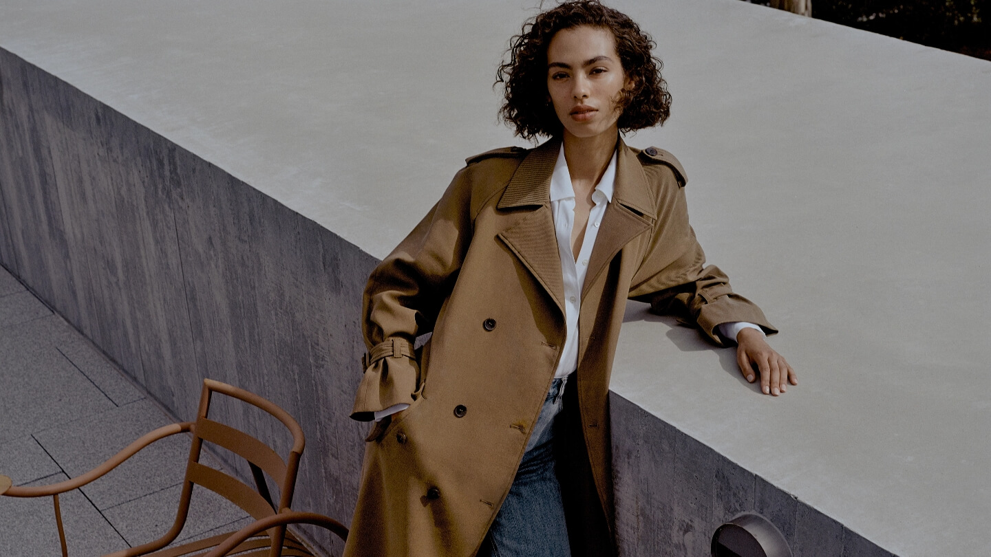 A woman wearing a tan trench coat stands on a ledge next to a concrete wall, accented by chairs. Fashion editorial style.