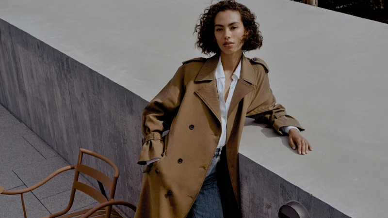 A woman wearing a tan trench coat stands on a ledge next to a concrete wall, accented by chairs. Fashion editorial style.