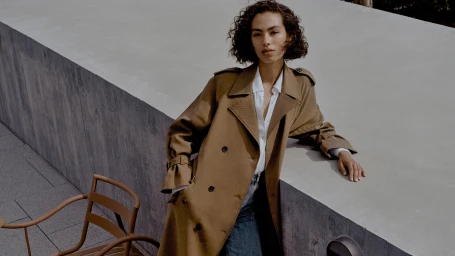 Model in a brown trench coat leaning against a concrete ledge outdoors