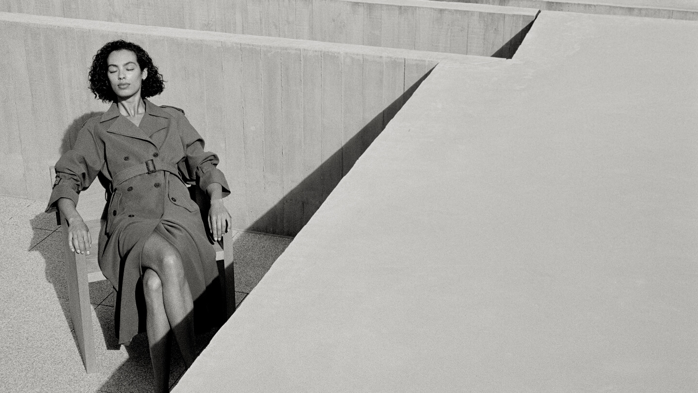 Elegant woman in a trench coat sits in sunlight against a concrete structure, creating a serene and fashionable black and white image.