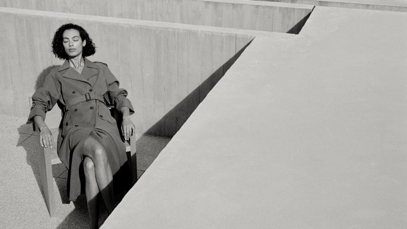Elegant woman in a trench coat sits in sunlight against a concrete structure, creating a serene and fashionable black and white image.