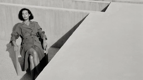 Woman wearing a belted trench coat seated outdoors against architectural concrete walls