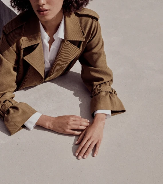 A woman wearing a tan trench coat and white shirt poses with her hands on a flat surface, photographed at eye level in soft lighting.