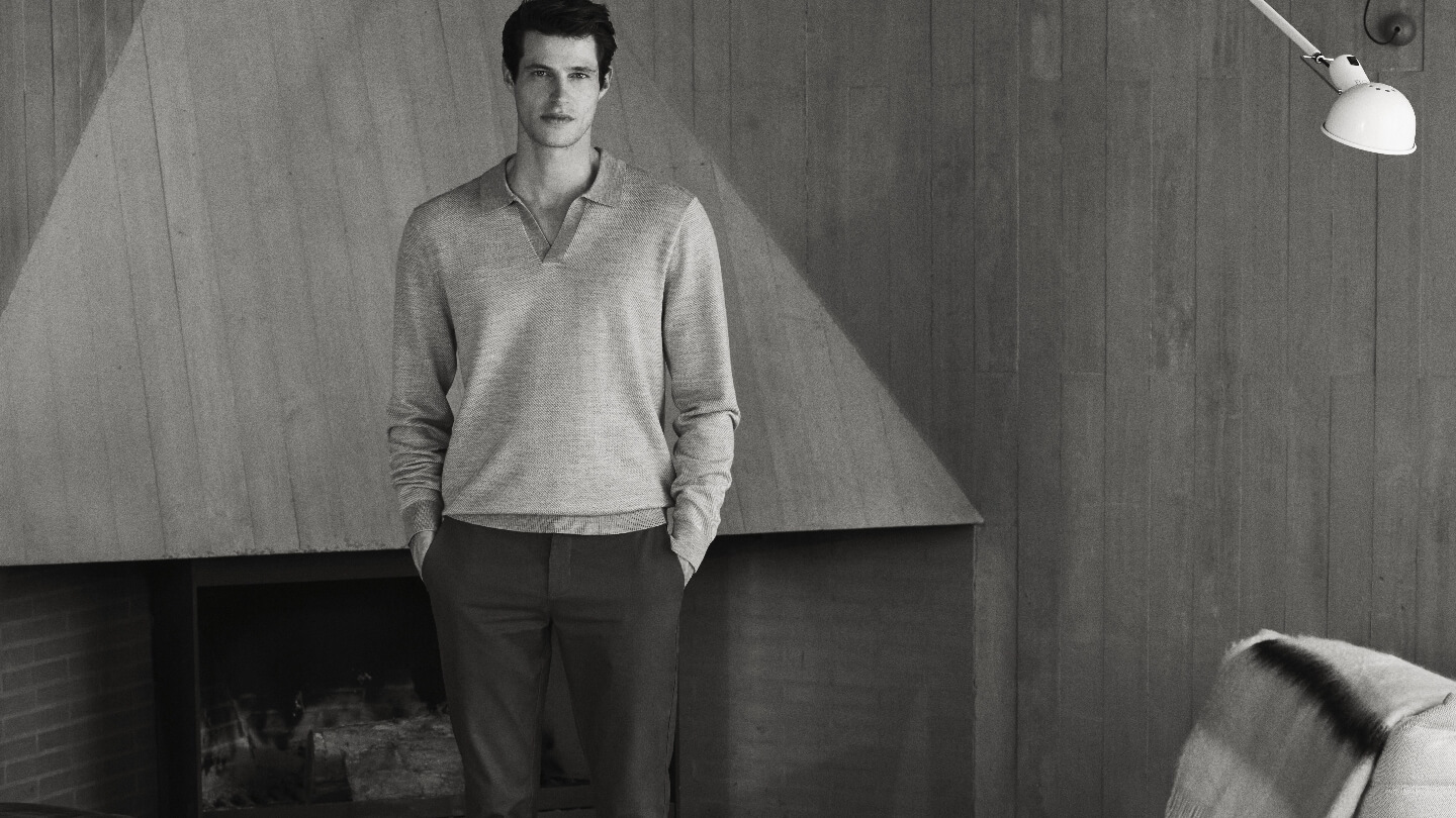 Monochrome portrait of a male model in a relaxed pose, wearing a grey collared shirt and dark trousers in a neutral indoor setting.
