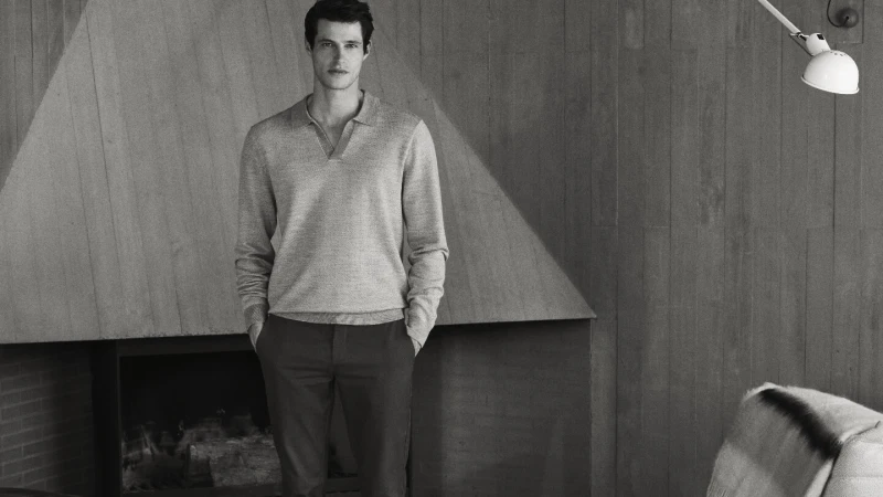 Monochrome portrait of a male model in a relaxed pose, wearing a grey collared shirt and dark trousers in a neutral indoor setting.