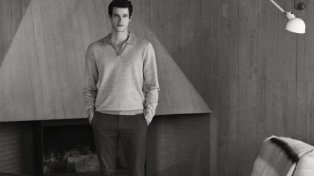 Monochrome portrait of a man standing in a modern living room wearing a long-sleeve knit polo shirt and trousers.
