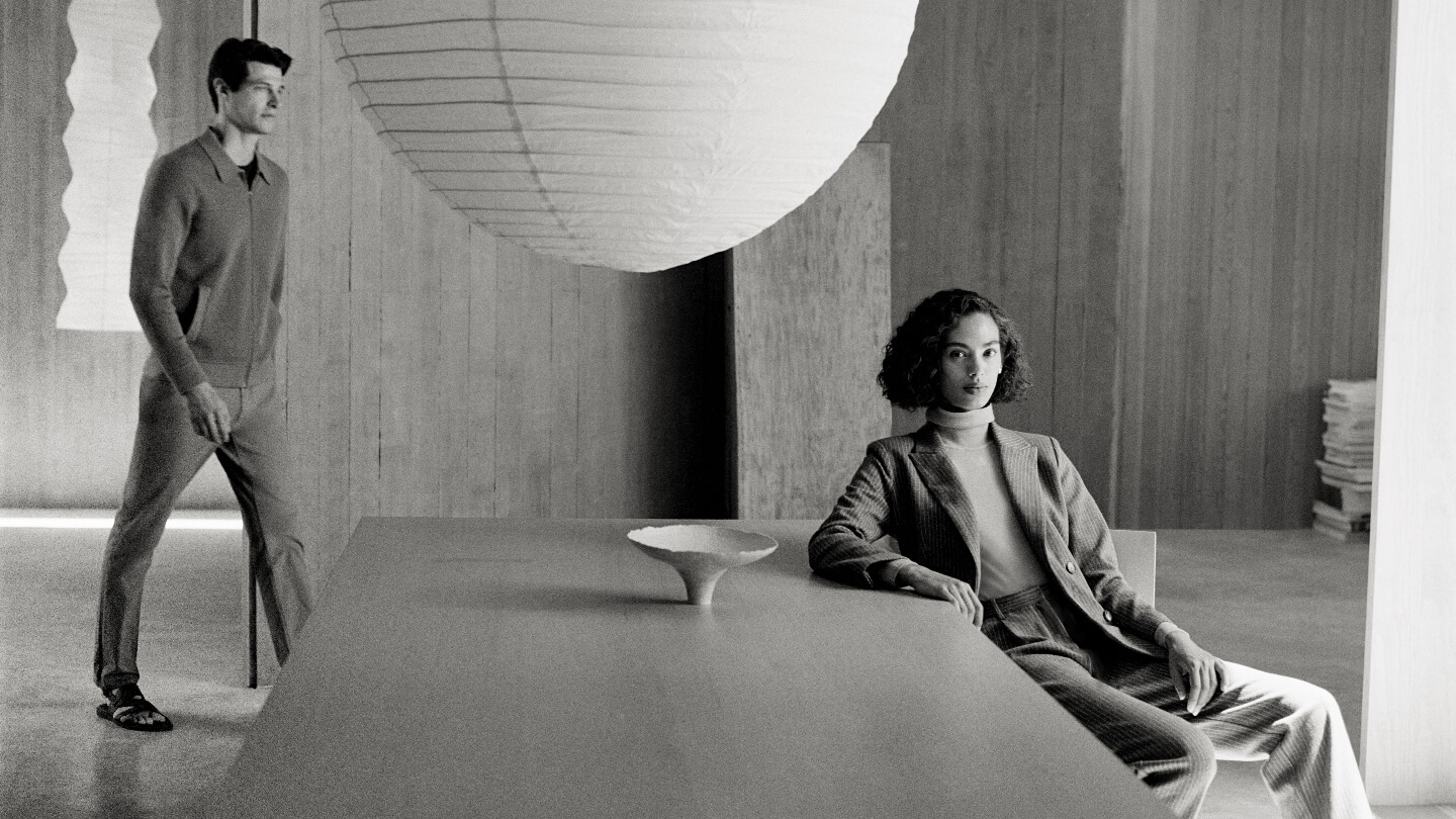 Monochrome fashion shot of a woman in a suit seated at a table with a bowl, and a man in a sweater and pants walking in the background.