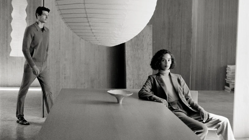 Monochrome fashion shot of a woman in a suit seated at a table with a bowl, and a man in a sweater and pants walking in the background.