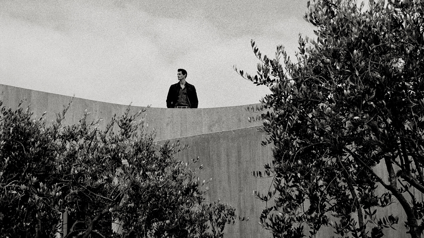 Monochrome portrait of a man standing on a concrete structure with trees in the foreground, gazing away in natural daylight.