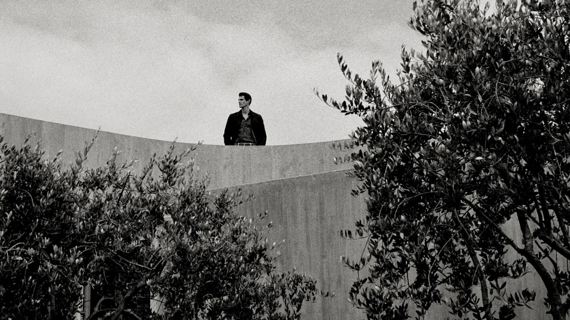 Monochrome portrait of a man standing on a concrete structure with trees in the foreground, gazing away in natural daylight.