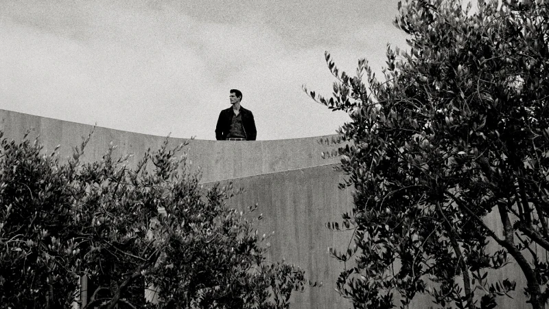 Monochrome portrait of a man standing on a concrete structure with trees in the foreground, gazing away in natural daylight.
