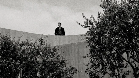 Monochrome image of a man standing atop a curved concrete wall framed by leafy trees under an overcast sky.