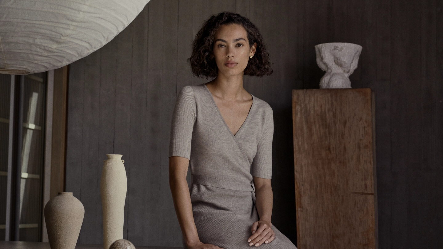 A woman with curly hair sits indoors wearing a beige wrap dress, looking forward; vases sit on a table with neutral wooden wall behind.