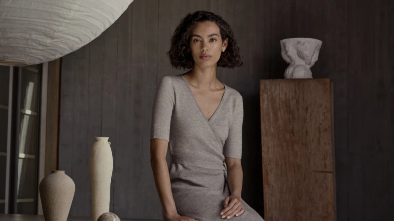A woman with curly hair sits indoors wearing a beige wrap dress, looking forward; vases sit on a table with neutral wooden wall behind.
