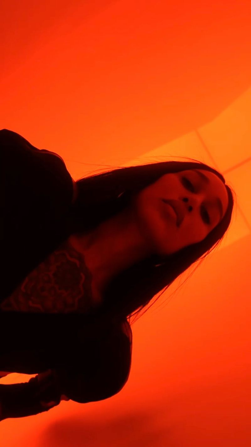 Portrait of an adult woman with long dark hair, wearing a dark top, posing with her eyes closed against an orange background.