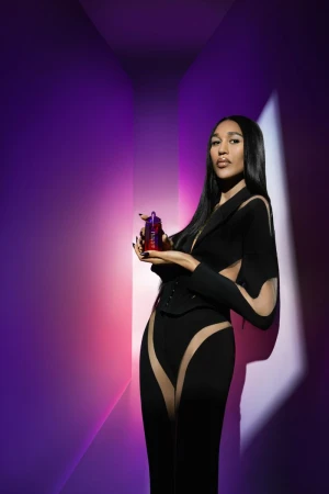 Elegant adult woman in a black cutout outfit poses with a purple perfume bottle against a vibrant purple and red gradient backdrop.