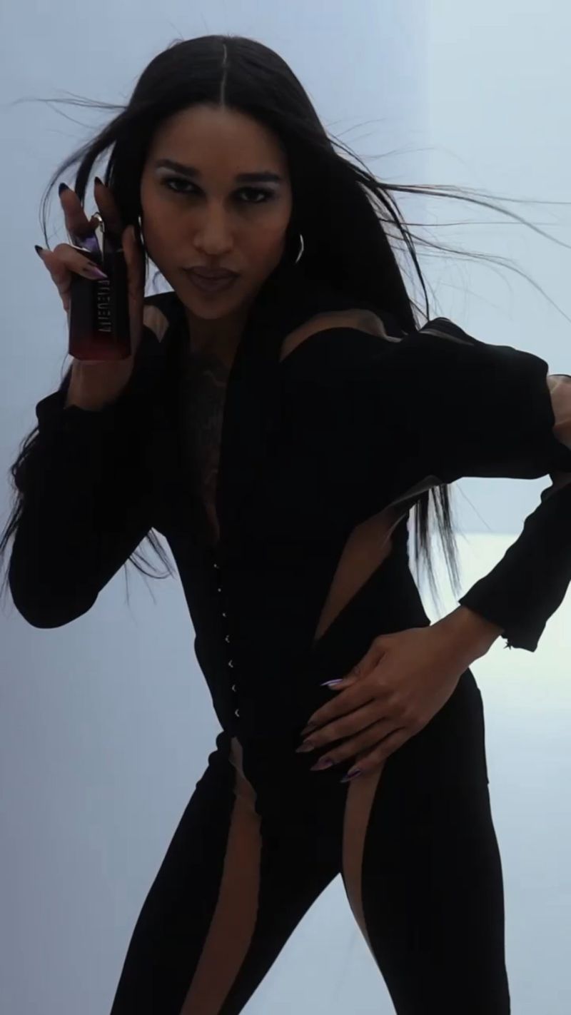 Adult woman in a black jumpsuit holds a cosmetic product. The woman's pose is dynamic, and the background is plain light-grey.